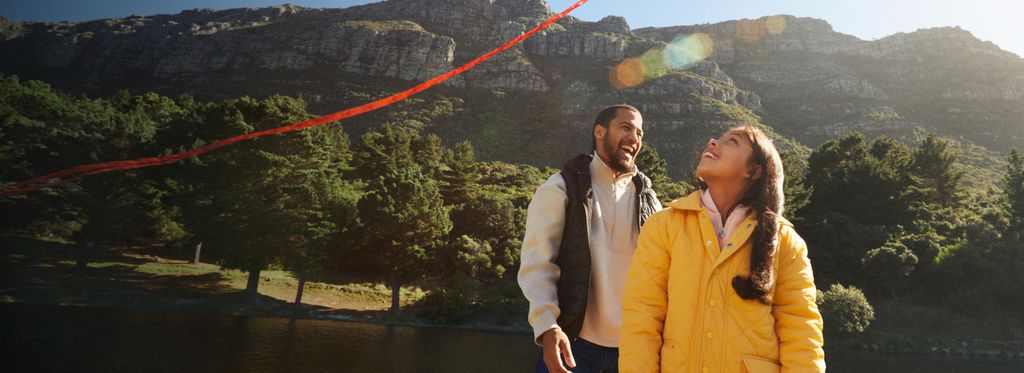 Man and two girls fly a kite by a river with mountains in the background