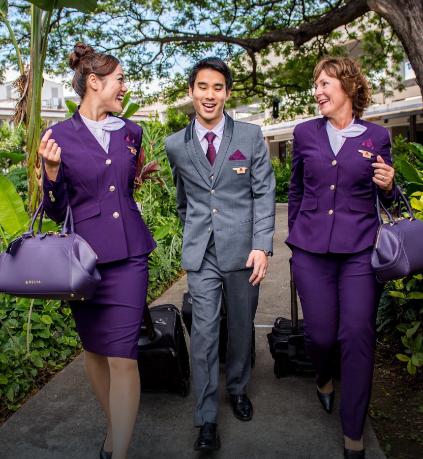 Delta flight attendants smiling while walking down flower and tree lined pathway with their luggage