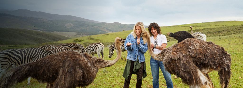 Two women feeding ostriches in a vast field with zebras and hills in the background