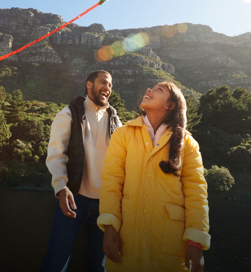 Man and two girls fly a kite by a river with mountains in the background