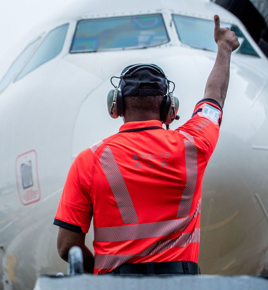 Delta ramp agent in orange uniform shirt gives thumbs up sign to pilots in cockpit of aircraft at gate