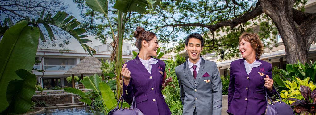 Delta flight attendants smiling while walking down flower and tree lined pathway with their luggage