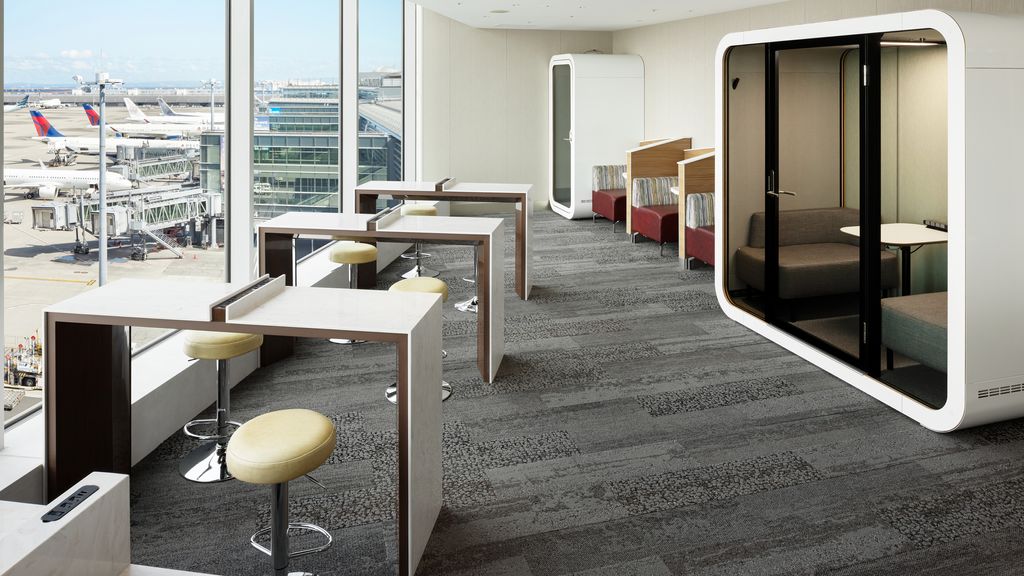 A modern airport lounge with a view of the tarmac and airplanes. The lounge features white tables with yellow stools and individual seating pods with private workspaces. Large windows provide natural light and a clear view of airport operations outside.
