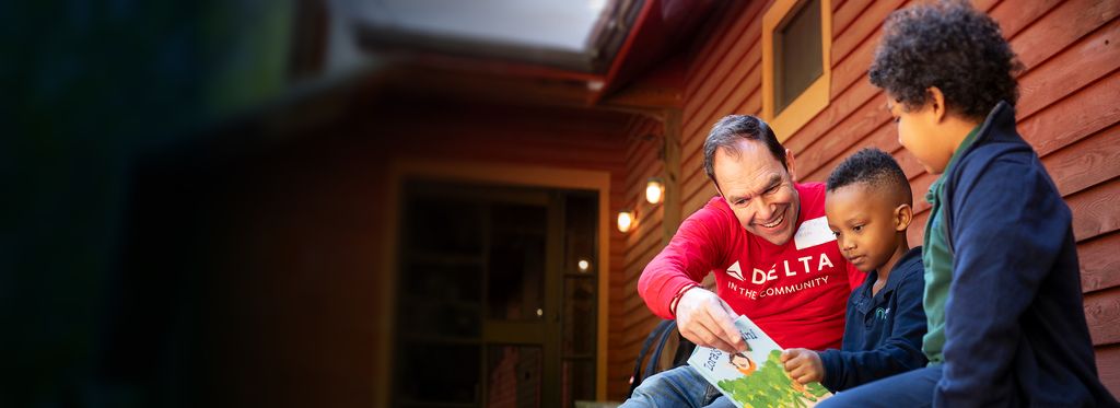 A Delta volunteer in a red Delta t-shirt is sitting on a bench, reading a book to two young people.