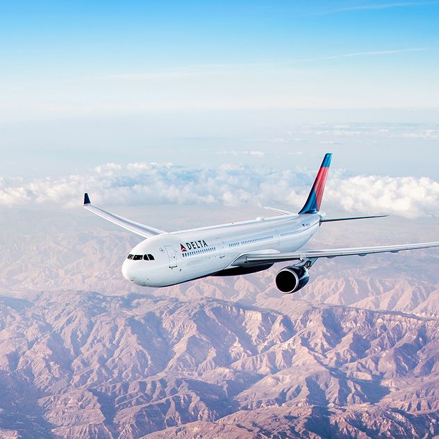 A Delta airplane flies over a vast, mountainous landscape with rugged terrain with clear skies and some distant clouds.