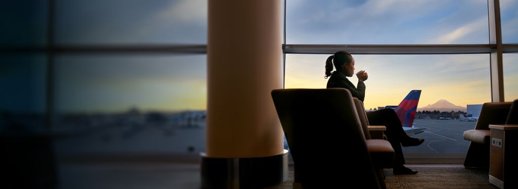 A person sitting in an airport lounge, looking out of a large window onto the tarmac. In the background, there is a mountain visible under the sunset sky.