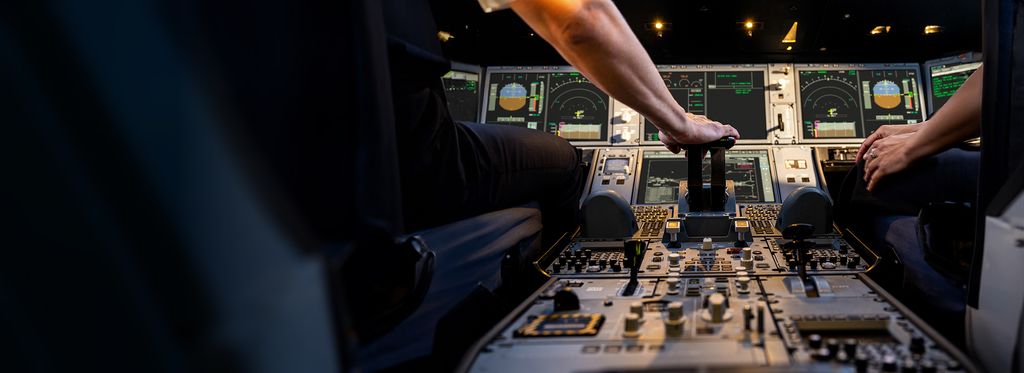A close up of the controls in the cockpit of an aircraft showing two pilots' hands. One pilot's hand is on the controls, and various instruments, buttons, and screens are visible.