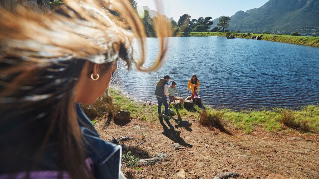 A group of people near a lake or pond with mountains and trees in the background. One person is in the foreground, while three others are closer to the water's edge.