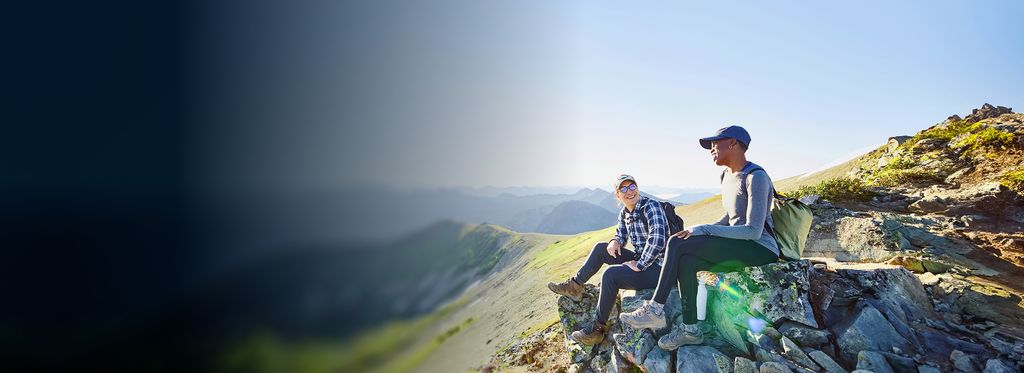 Two people sitting on a rocky ledge overlooking a mountainous landscape. The sky is clear and bright, and the terrain below includes green valleys and distant mountain ranges.