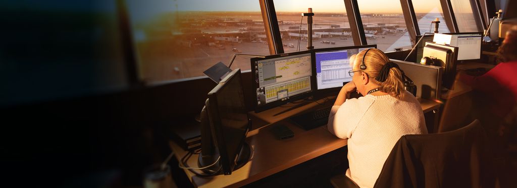 A woman sits at a desk with two computer monitors in an air traffic control tower.