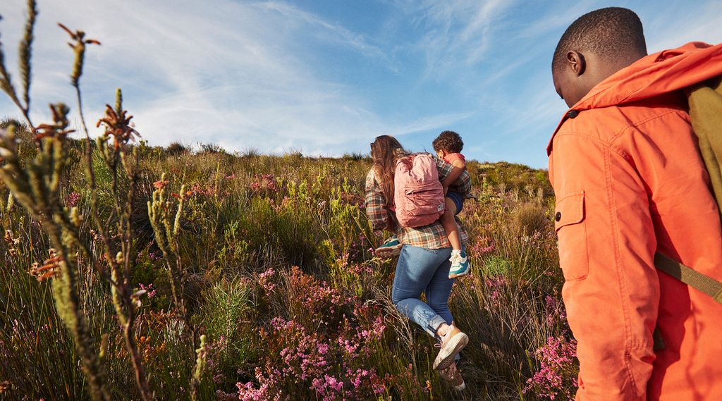 A group of people hiking up a hill covered in wildflowers and vegetation. One person is carrying a child on their back, while another person wearing an orange jacket follows behind.