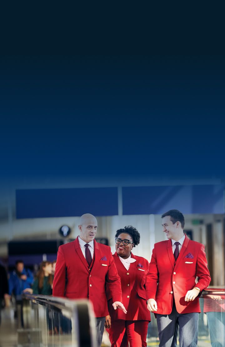 Three Delta customer service employees wearing red uniforms, walking on a moving walkway in an airport.