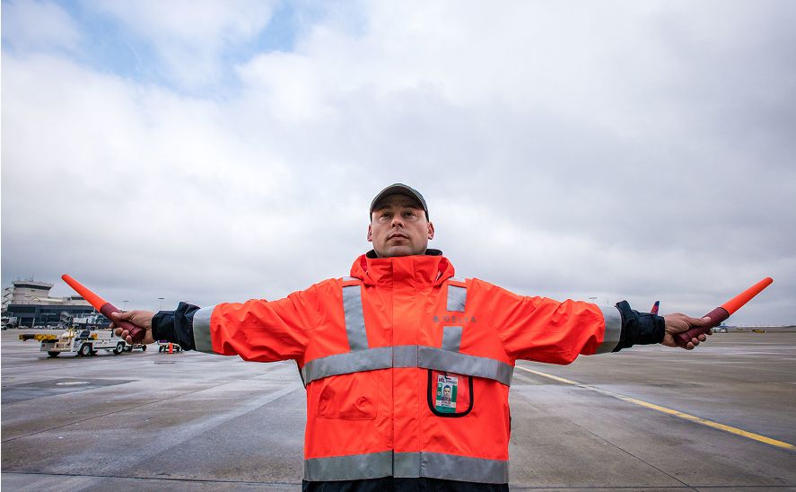 A Delta ramp employee on the airport tarmac, wearing an orange high-visibility jacket and holding two orange batons with arms extended outward.