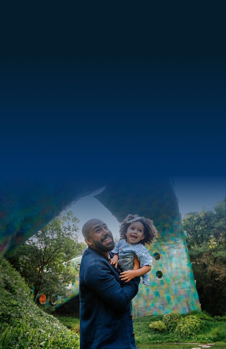 A man holding a child outdoors with greenery and a colorful structure in the background.