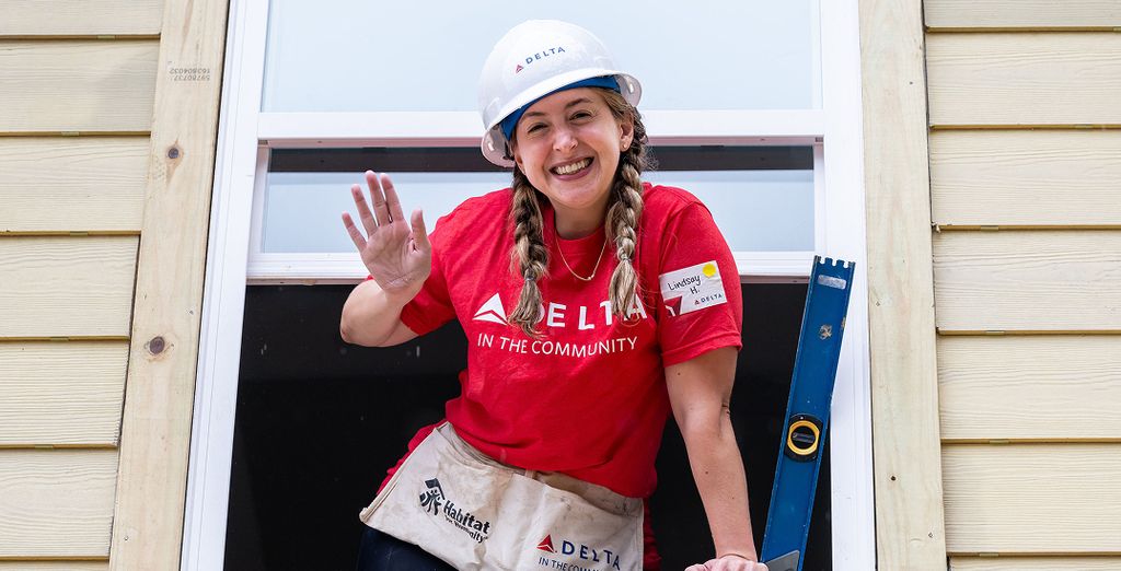 A person in a white hard hat and red 'DELTA IN THE COMMUNITY' shirt volunteers at a Habitat for Humanity build.