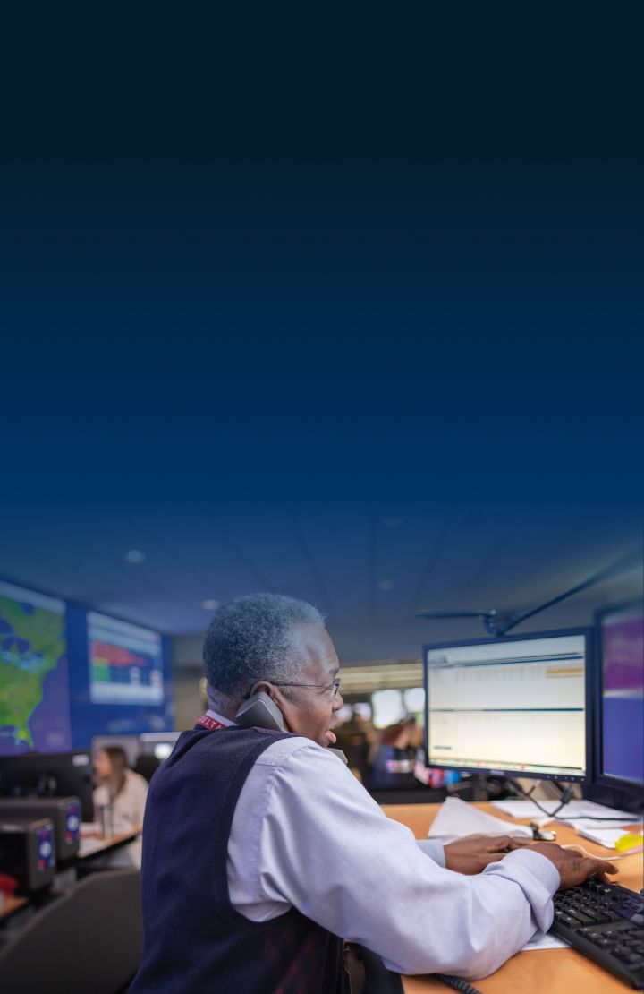 A Delta employee works at a desk with two computer monitors in a control room, wearing a white shirt and dark vest.