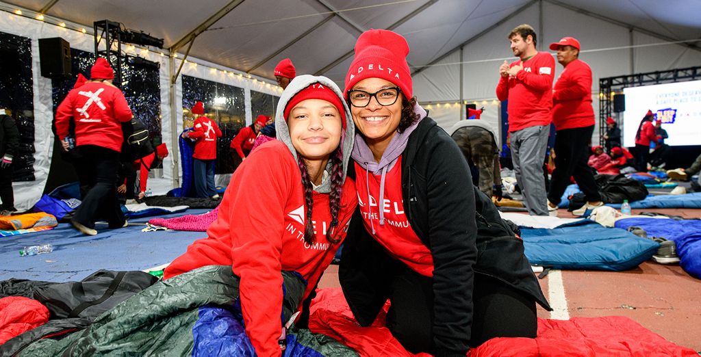 A group of people inside a large tent wearing red Delta shirts and hats surrounded by blankets and sleeping bags.