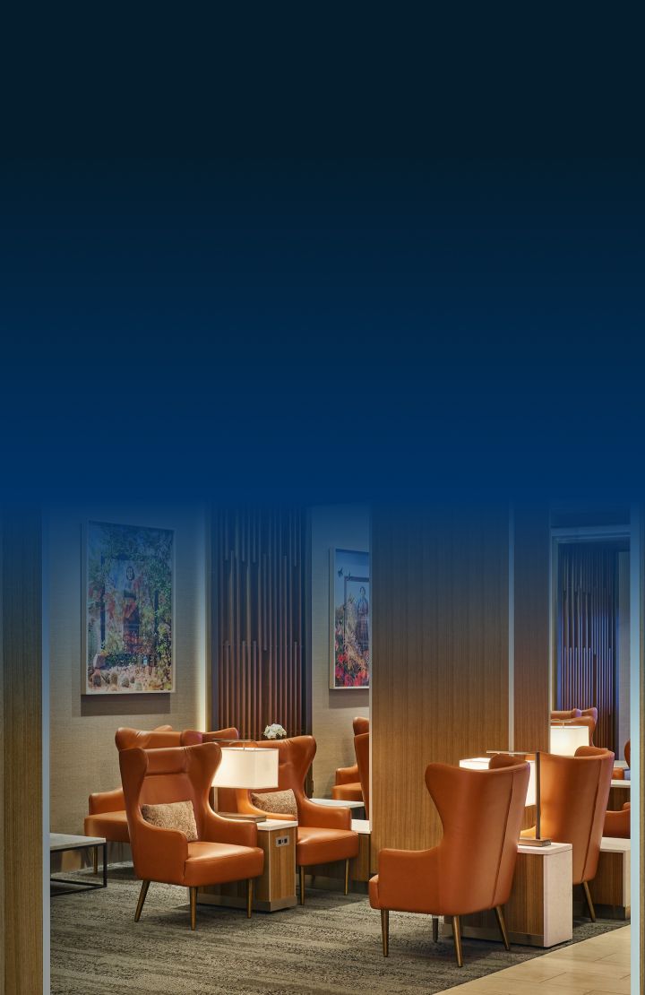 A Delta Sky Club lounge with warm lighting, leather armchairs, wood paneling, and elegant decor, including artwork and a statement chandelier.