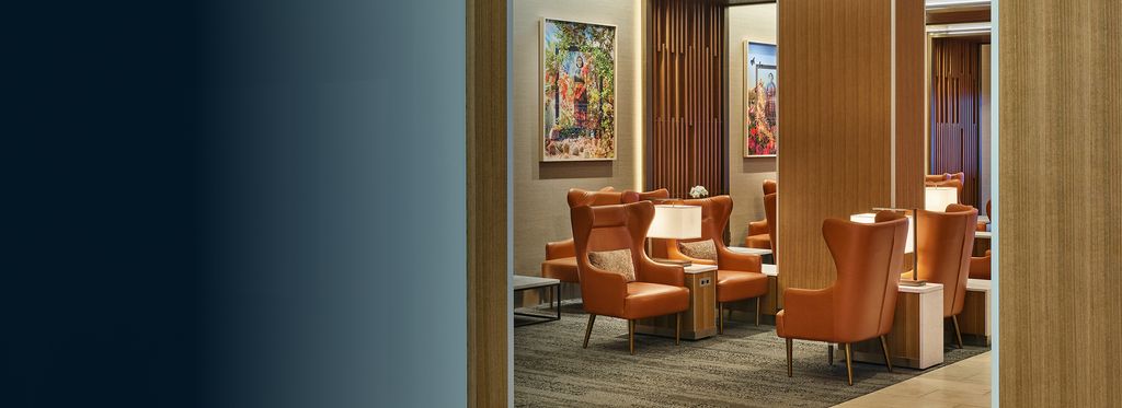 A Delta Sky Club lounge with warm lighting, leather armchairs, wood paneling, and elegant decor, including artwork and a statement chandelier.