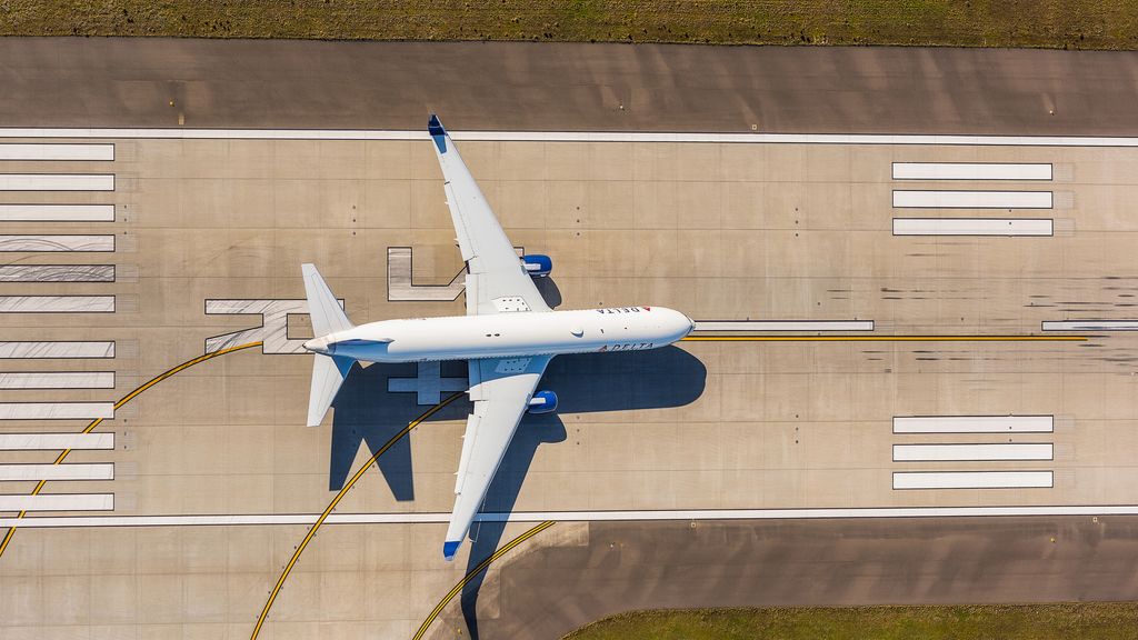 An aerial view of a Delta airplane centered on a runway with white markings and flanked by grassy areas on both sides.