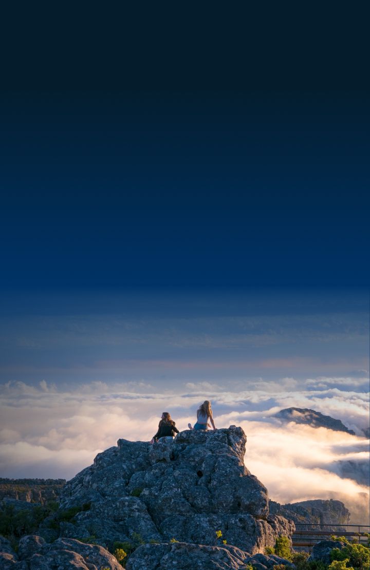 Two people are sitting on a rocky outcrop, overlooking a mountainous landscape with clouds rolling over the peaks.