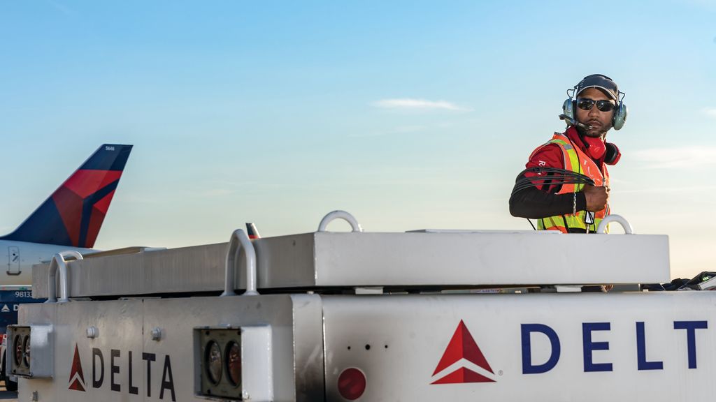 A Delta ramp agent on ground support equipment on the tarmac, wearing a headset and a high-visibility vest