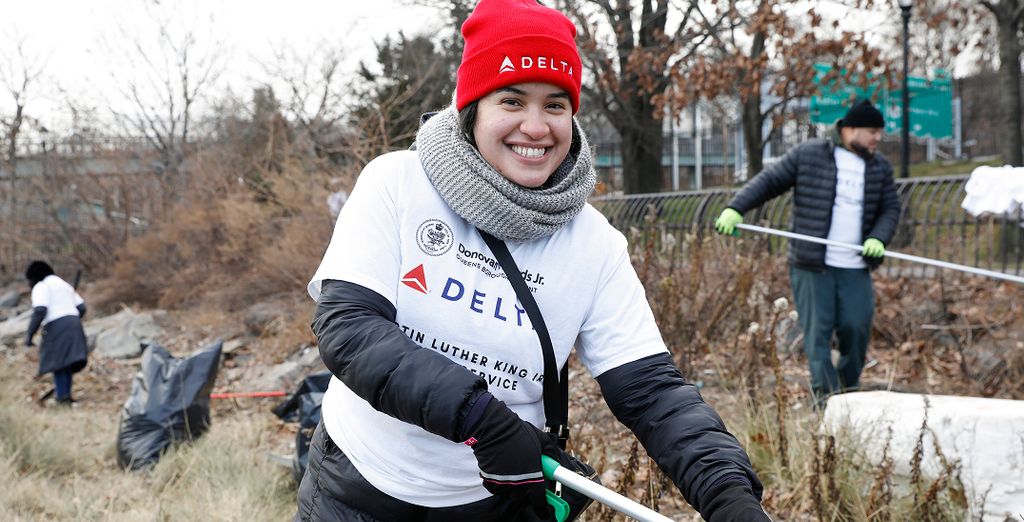 A person wearing a red hat and a white DELTA t-shirt holding a trash picker during an outdoor cleanup activity.