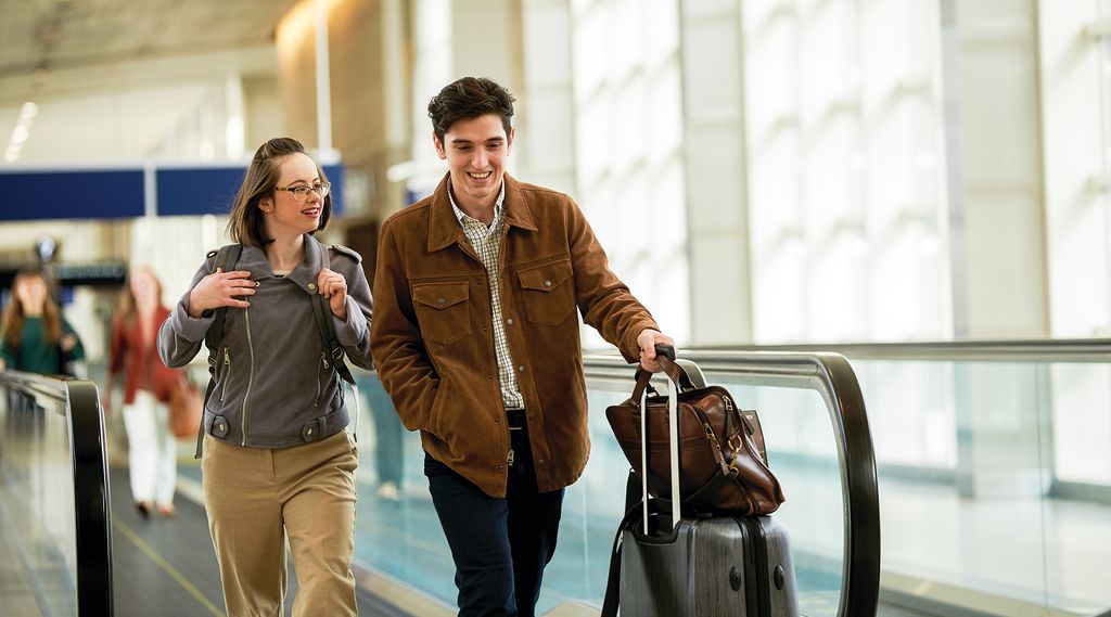 Two people on a moving walkway in an airport. One is pulling a suitcase and holding a bag and the other is wearing a backpack and holding onto the straps.