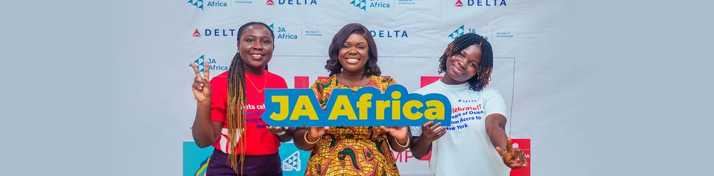 Three ladies standing in front of a backdrop with the logos of 'JA Africa' and 'Delta'. They are holding a sign that reads 'JA Africa'.