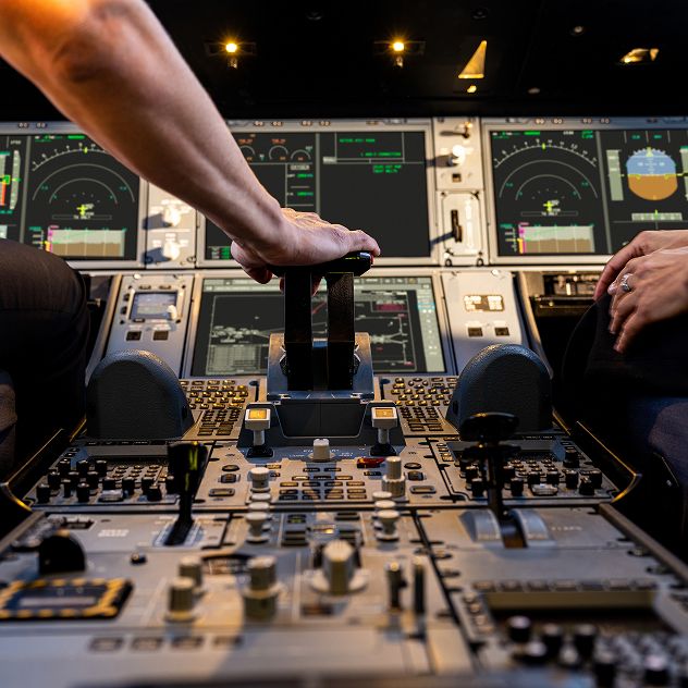 A close up of the controls in the cockpit of an aircraft showing two pilots' hands. One pilot's hand is on the controls, and various instruments, buttons, and screens are visible.