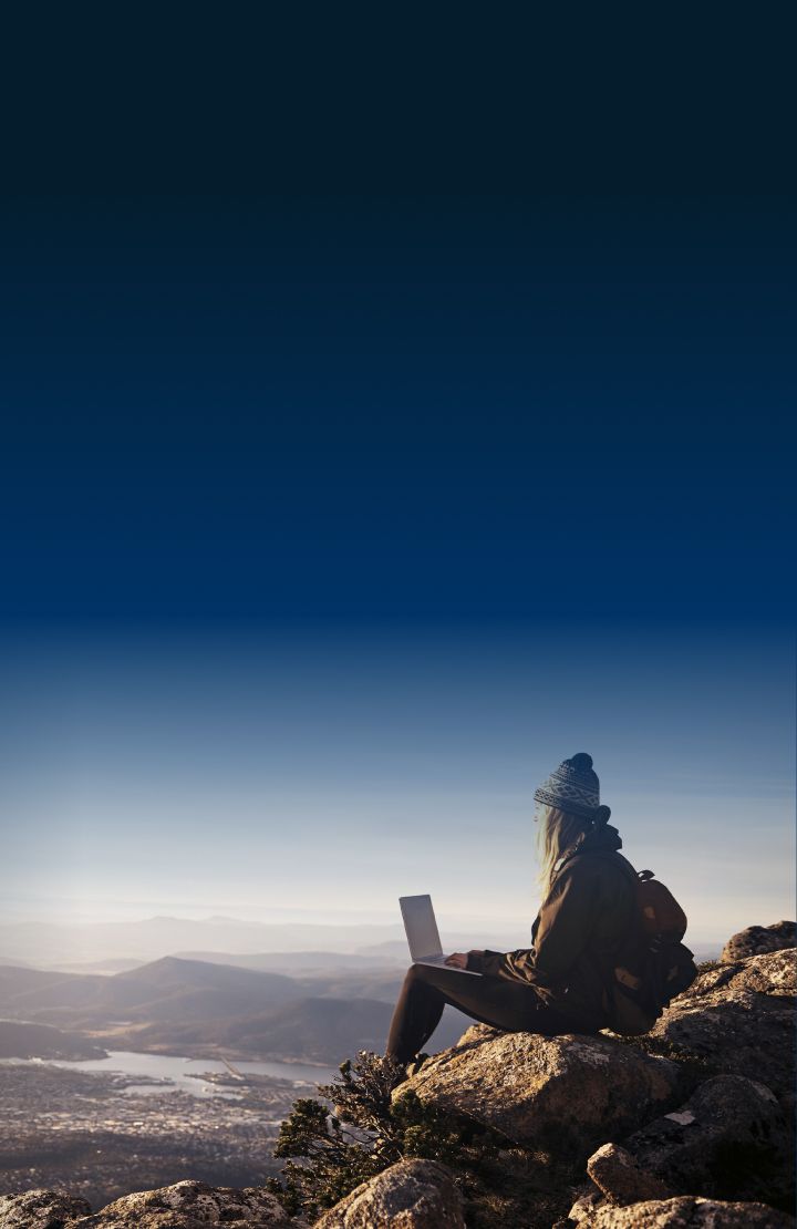 A person sits on a rocky cliff at sunrise, overlooking mountains and valleys, using a laptop.