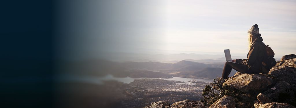 A person sits on a rocky cliff at sunrise, overlooking mountains and valleys, using a laptop.