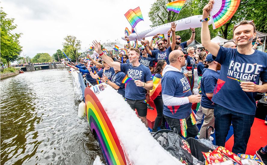A group of people wearing blue 'PRIDE IN FLIGHT' T-shirts holding rainbow flags and accessories celebrate on a rainbow-designed float along a waterway.