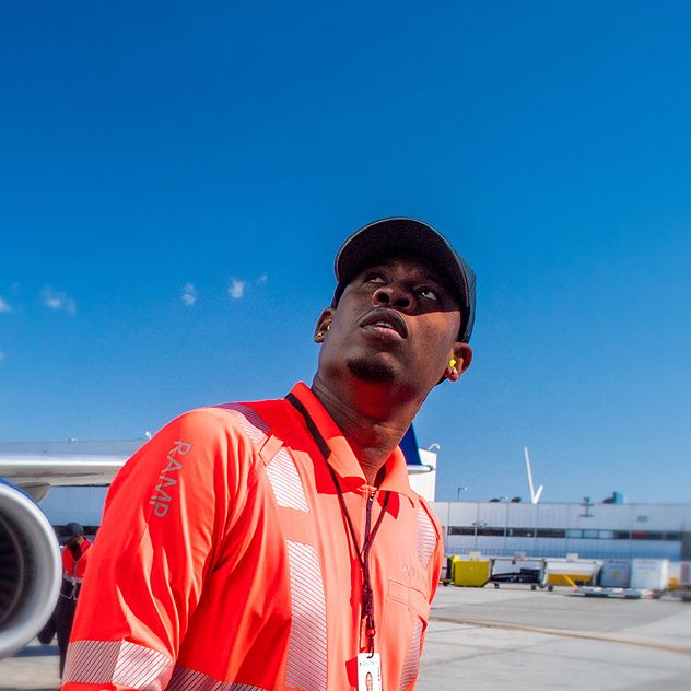 A Delta ground employee in an orange high-visibility vest holding two orange marshalling wands walks near a large airplane on an the tarmac.