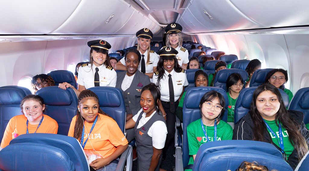 Delta airplane cabin with young female passengers seated and several female pilots and flight attendants standing in the aisle