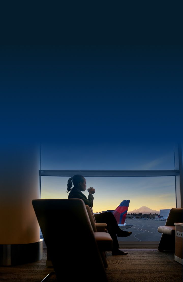 A person sitting in an airport lounge, looking out of a large window onto the tarmac. In the background, there is a mountain visible under the sunset sky.