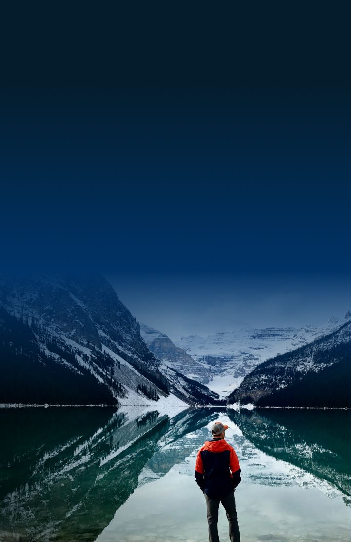 A person standing by a calm lake reflecting snow-capped mountains.