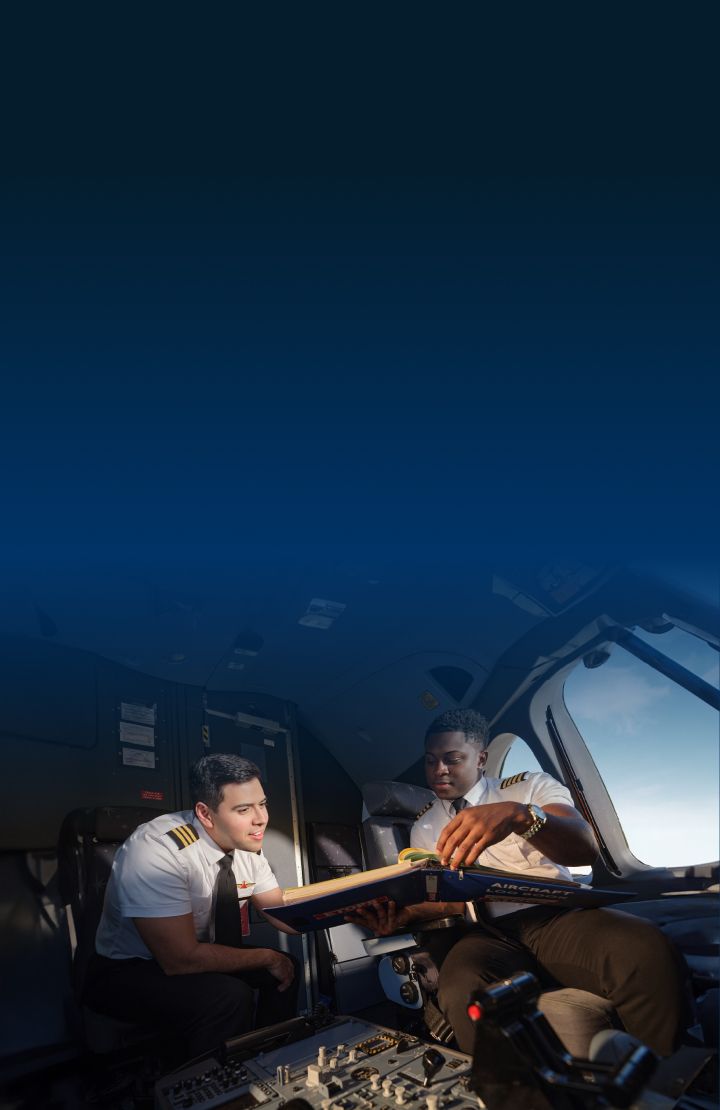 Two pilots in the cockpit. One holds a manual, while the other leans over to look at it. Various controls and instruments are visible.
