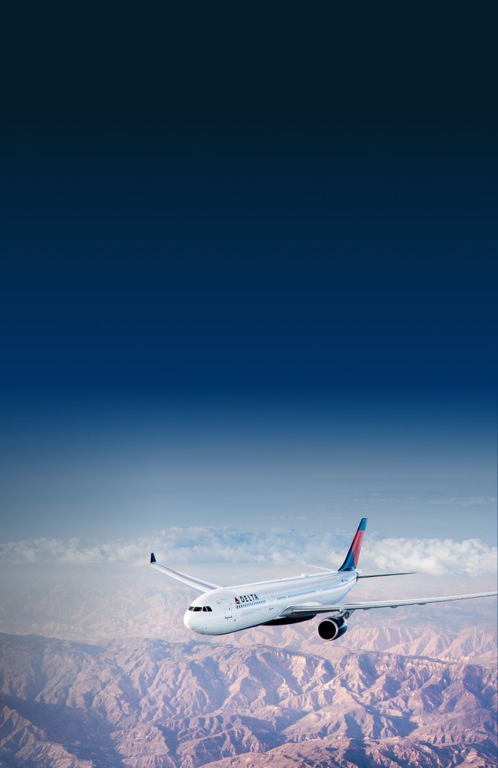 A Delta airplane flies over a vast, mountainous landscape with rugged terrain with clear skies and some distant clouds.