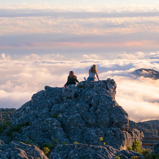 Two people are sitting on a rocky outcrop, overlooking a mountainous landscape with clouds rolling over the peaks.