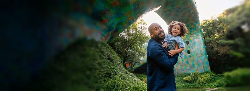 A man holding a child outdoors with greenery and a colorful structure in the background.