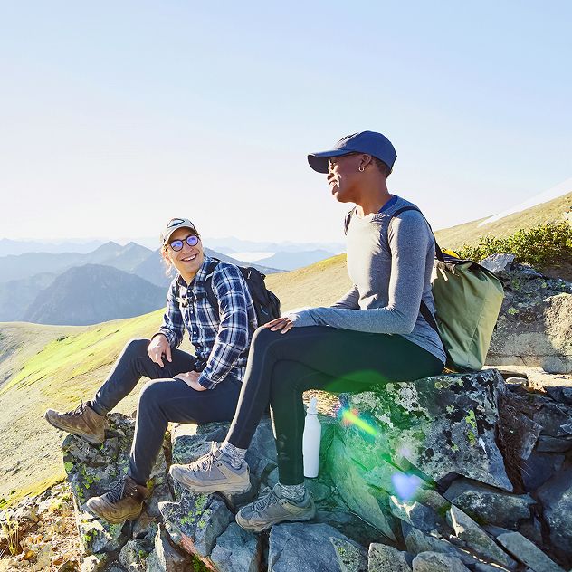 Two people sitting on a rocky ledge overlooking a mountainous landscape. The sky is clear and bright, and the terrain below includes green valleys and distant mountain ranges.