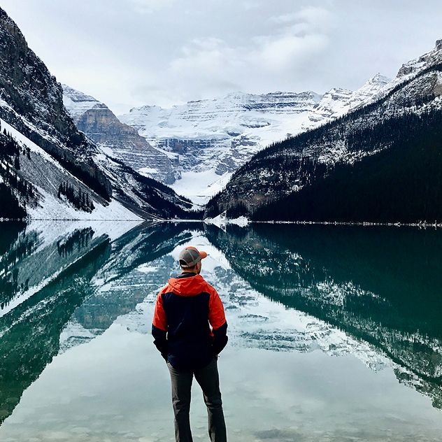 A person standing by a calm lake reflecting snow-capped mountains.