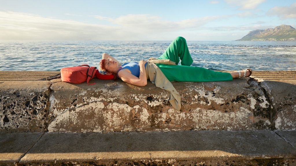 A person wearing green pants and a blue top lying on a concrete wall by the sea with a red bag under their head.