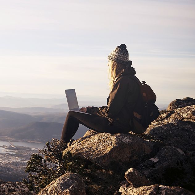 A person sits on a rocky cliff at sunrise, overlooking mountains and valleys, using a laptop.