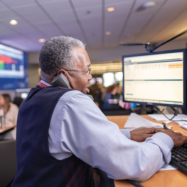 A Delta employee works at a desk with two computer monitors in a control room, wearing a white shirt and dark vest.