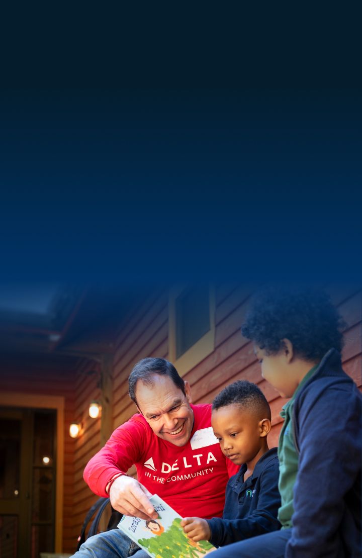 A Delta volunteer in a red Delta t-shirt is sitting on a bench, reading a book to two young people.