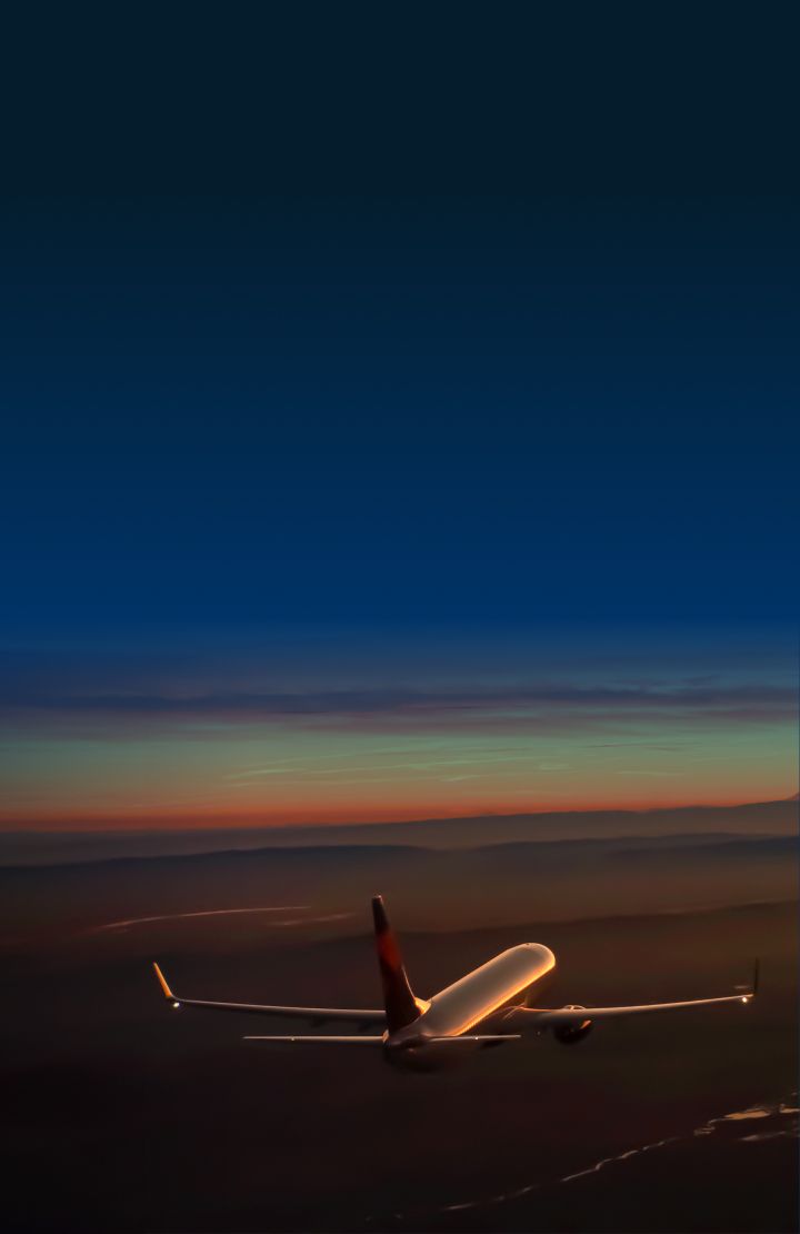 A Delta airplane flies above dark terrain at sunset, with the sky displaying orange and yellow hues.