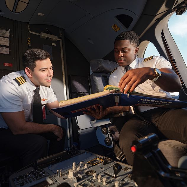 Two pilots in the cockpit. One holds a manual, while the other leans over to look at it. Various controls and instruments are visible.