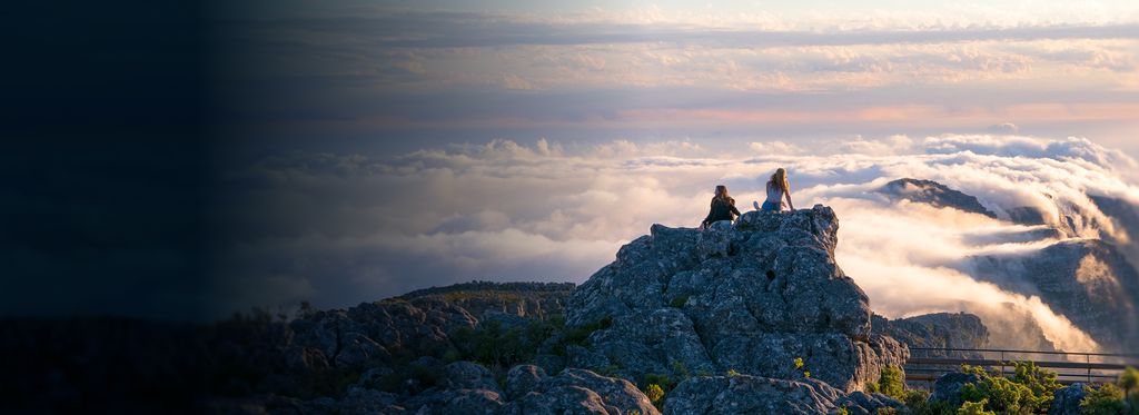 Two people are sitting on a rocky outcrop, overlooking a mountainous landscape with clouds rolling over the peaks.